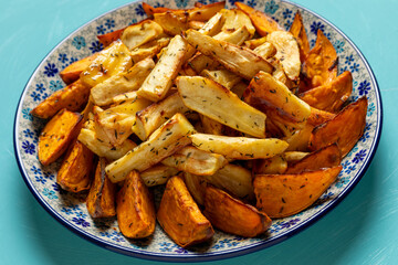 A platter with baked root vegetables: parsnip in the middle surrounded by sweet potato pieces. Plain blue background.