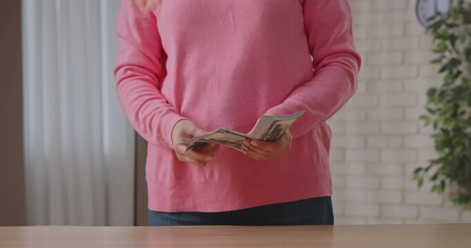 A woman in a pink sweater is holding and counting a stack of hundred dollar bills at a table indoors. Slow motion.