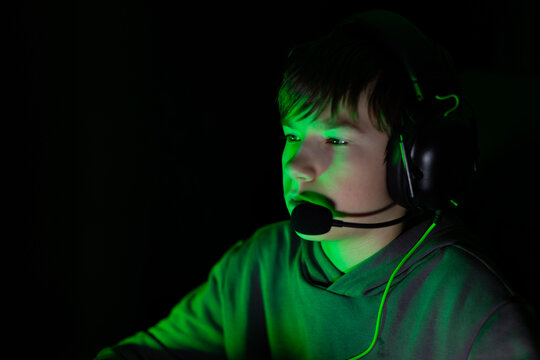 Young male gamer wearing a headset, focused on screen illuminated by green light, showcasing intense concentration and engagement in online gaming experience - Powered by Adobe
