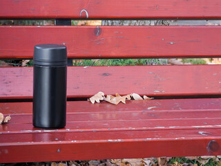 Black thermos mug standing on a vibrant red wooden park bench, surrounded by scattered autumn leaves, evoking warmth and comfort during outdoor relaxation