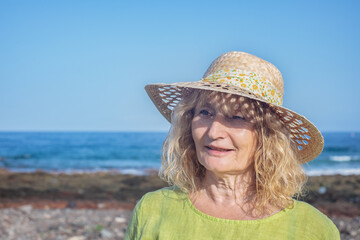 Portrait of smiling middle aged blonde woman with straw hat standing outdoors at the sea beach.  Vacation, freedom, concept. Horizon over water