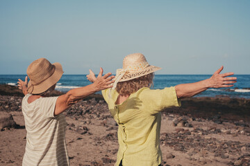 Rear view of two caucasian women with outstretched arms standing at the beach face the sea looking the horizon. Freedom, comfort and friendship concept