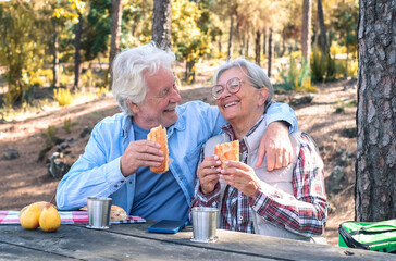 Happy senior couple enjoying a picnic outdoors in a sunny forest. The elderly man and woman are sitting at a wooden table, laughing sharing food and drinks. Positive retired lifestyle in nature