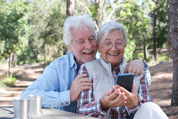 Happy senior couple enjoying a picnic outdoors in a sunny forest using smartphone. Elderly man and woman sitting at a wooden table smiling sharing food and drinks. Positive retired lifestyle in nature