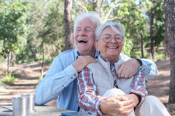 Cheerful senior couple enjoying a picnic outdoors in a sunny day in the forest. Elderly man and woman sitting at a wooden table laughing with fun. Positive retired lifestyle in nature