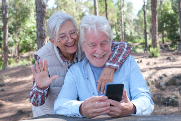 Happy senior couple enjoying a picnic outdoors in a sunny forest using smartphone. Elderly man and woman sitting at a wooden table smiling sharing food and drinks. Positive retired lifestyle in nature