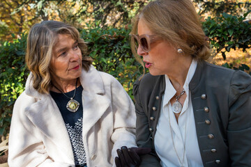 Two elegant women share a heartfelt conversation in a vibrant autumn garden filled with colorful foliage