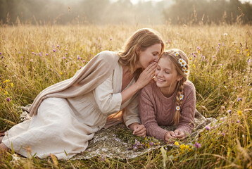 A mother tenderly whispers a secret to her smiling daughter while relaxing together in a sunny field. Concept of family love, trust, and connection.