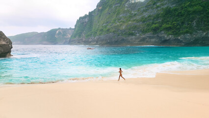 Woman Walking Along Turquoise Shoreline On Remote Tropical Beach With Cliffs