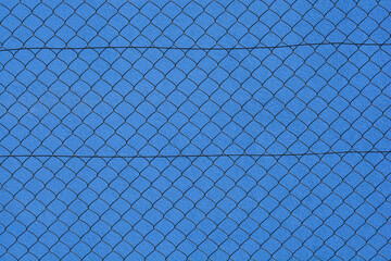 A front view of a diamond pattern wire mesh fence stretching across a textured blue plaster wall background