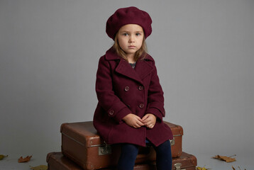 A stylish little girl in a retro coat and beret sits on vintage suitcases. Autumn travel, childhood, and kids' fashion concept.
