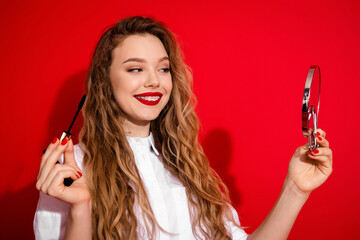 Young woman holding a makeup brush and hand mirror while smiling in a red background showcasing...