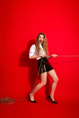 Stylish young woman pulling rope on red background with cheerful expression, wearing chic outfit...