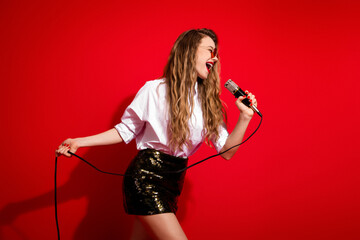 Excited young woman singing into a microphone on a vibrant red background, expressing joy and energy with stylish attire