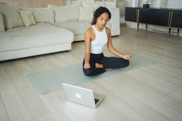Yoga mindfulness meditation online. African woman practicing yoga with online lessons in laptop at...
