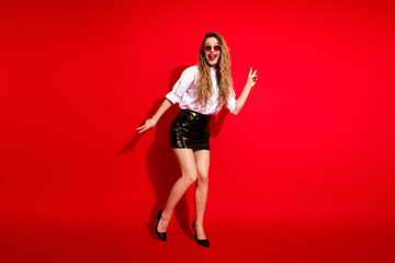 Stylish young woman in a fashionable outfit posing playfully against a vibrant red background,...