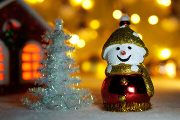 Against a blurred background with bright lights and a house, a snowman with a white Christmas tree