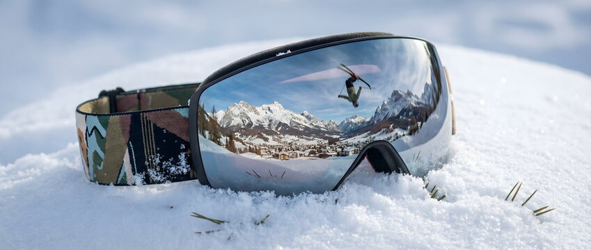 Ski goggles on snow with reflection of freestyle skier jumping in mountains. Winter sports equipment and alpine resort vacation concept.