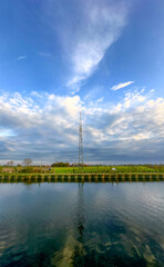 A tranquil scene featuring a towering communication structure reflected in calm water under a picturesque sky.