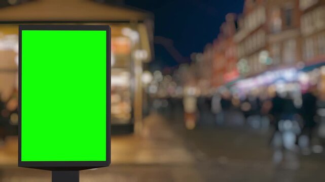 Green screen billboard standing on a busy city street at night with blurred lights and people passing by. Perfect for advertisement