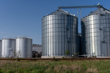Large silos are located in a field with green grass. The blue sky shows no clouds. The facility is used for storing grain and other agricultural products