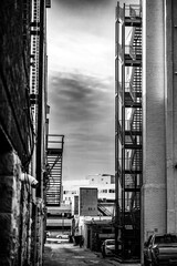 Alley view between buildings featuring a metal fire escape and distant Wells Fargo sign in the background during daylight