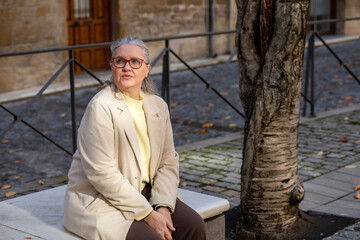 Elegant woman seated thoughtfully on a stone bench in a charming old town square during autumn