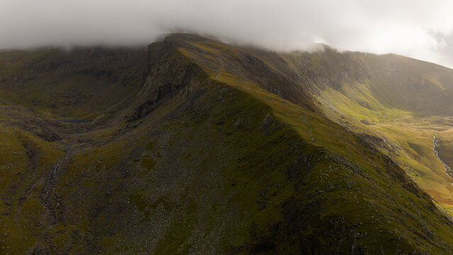 Aerial view of craggy peaks rise dramatically, shrouded in mist, their steep slopes a tapestry of green and brown, contrasting sharply with the cloudy sky, Snowdonia, Wales, United Kingdom.