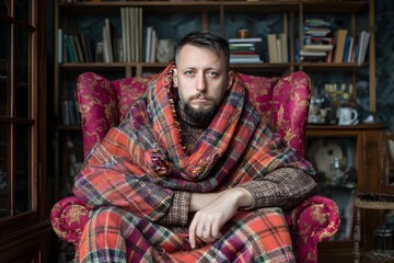 Fototapeta premium Man sitting on armchair wrapped in plaid blanket in cozy winter cabin
