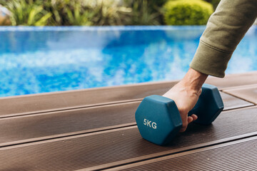 Individual lifting a blue dumbbell weighing 5kg on a wooden deck beside a swimming pool, showcasing fitness and strength training in a vibrant outdoor setting