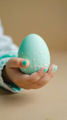 A child's hand holds a pastel blue Easter egg decorated with white dots. The background is neutral, emphasizing the egg's vibrant color.