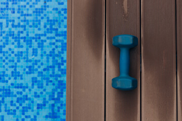 Blue dumbbell resting on wooden deck beside a shimmering swimming pool, reflecting sunlight, creating a vibrant and inviting atmosphere for fitness enthusiasts and relaxation seekers