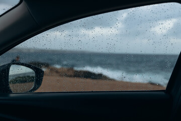 Rain droplets on car window create a blurred view of the ocean waves crashing against the rocky shore, capturing a serene coastal atmosphere during a rainy day