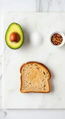Flat lay of avocado toast ingredients including fresh avocado, toasted bread, sea salt, and chili flakes on a white marble board.