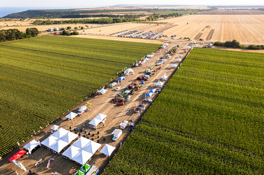 Aerial view of a bustling agricultural fair with rows of tents and machinery dividing fields of vibrant green and golden crops under a clear sky, Silistra, Bulgaria.