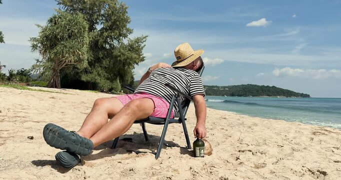 A drunk man with a bottle of rum relaxes on a sandy beach by the sea. A drunk man sleeps on the beach, a bottle of alcohol nearby. A vacation by the sea or ocean in a tropical country. Slow motion