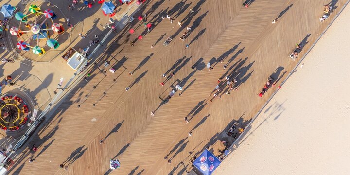 Aerial view of long shadows stretch across the wooden planks of a bustling boardwalk near the beach, casting a warm hue over Coney Island, Brooklyn, New York, United States.