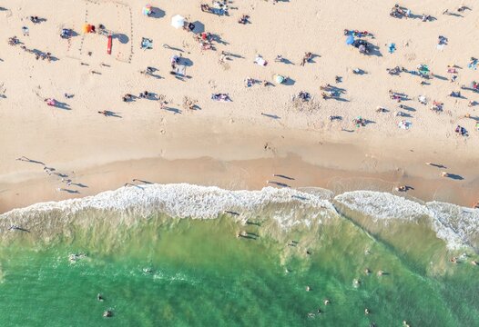 Aerial view of sunbathers dotted across the warm sand where the turquoise waves crash, creating a striking contrast of color and texture, Brooklyn, New York, United States.