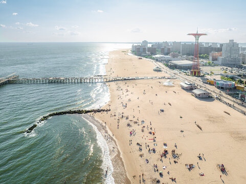 Aerial view of sun-kissed sands meet the rhythmic ocean waves, with the iconic Parachute Jump piercing the skyline above the bustling Coney Island boardwalk, Brooklyn, New York, United States.