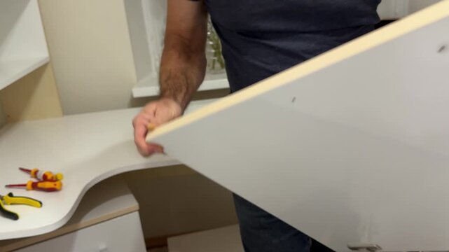 Professional furniture assembler carefully securing white shelf to mounting support, systematically tightening screws with precision screwdriver for stable furniture installation. High quality 4k