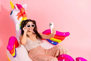 Fotobehang Diamanten Young woman in glitter dress relaxing on a colorful unicorn floatie taking a selfie with a smartphone in a pink studio background  © deagreez