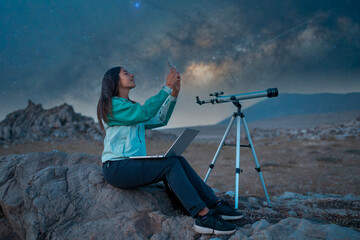 Young woman observing night sky with smartphone, laptop, and telescope on a rocky landscape