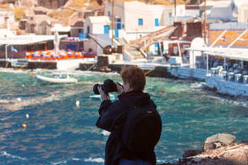 Tourist with camera photographing the waterfront and tavernas in Ammoudi Bay below Oia village on Santorini island, Greece.
