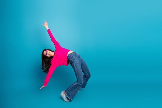 Young woman in pink sweater leans and raises arm in a playful dynamic pose against a blue studio backdrop for a fun fashion lifestyle stock image