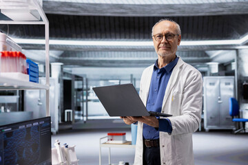 Fototapeta premium Portrait of male researcher standing in modern laboratory with white coat, working with multiple professional tools. Man in lab using laptop for biotechnology research and discovery.
