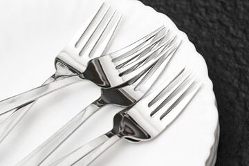 Four stainless steel forks on white plate. Modern table setting on black stone background. Cutlery...