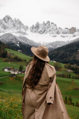 Woman in hat and trench coat looking at snow-capped mountains and green valley