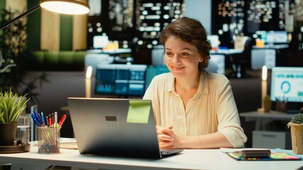 Professional woman sits in front of laptop during a teleconference, engaged in digital discussion. Interacting with participants, demonstrating adaptability in networking session. Camera B.