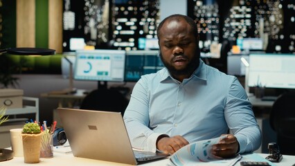 African american employee working at night proofreading web information, writing important data on laptop. Creating a summary and designing strategy to align with enterprise targets.