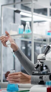 Vertical video Researcher pipetting reactant into blue solution for purity analysis. Female cytotechnologist adding liquid agent to test tube to observe chemical reaction properties, camera B
